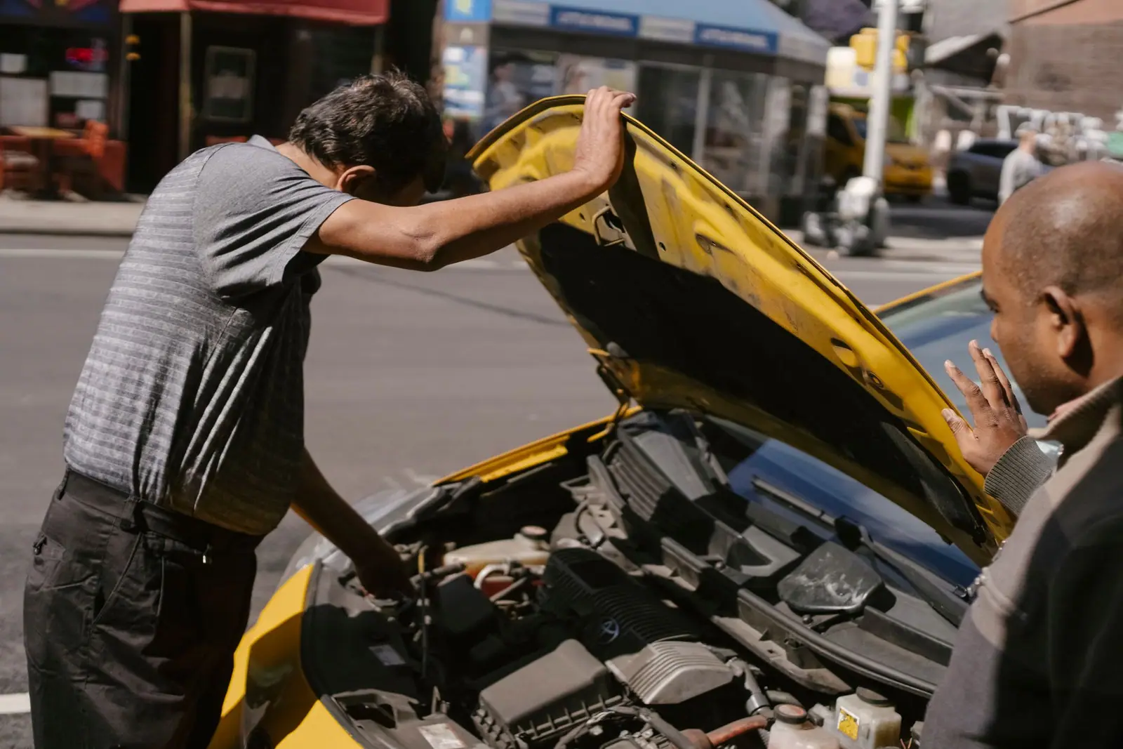 Two people looking into a car engine compartment beneath a raised yellow hood