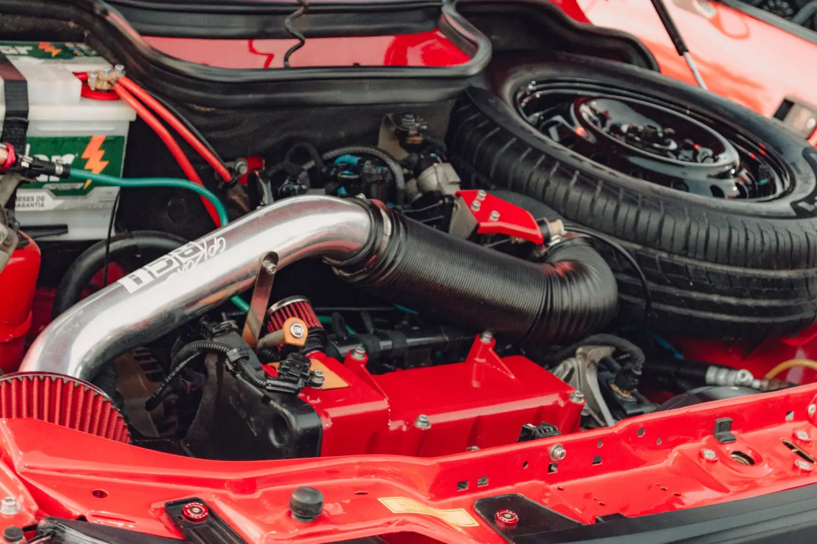 A red car engine compartment with a battery, metal tubing and a spare wheel visible