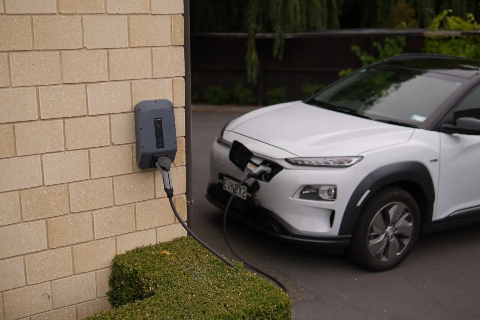 A white electric car charging from a wall-mounted unit beside a house