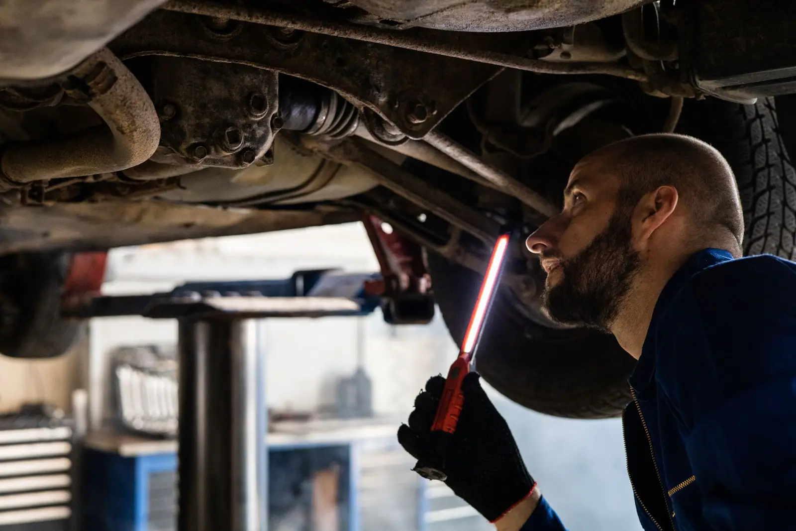 A mechanic using a work light to inspect the underside of a raised car